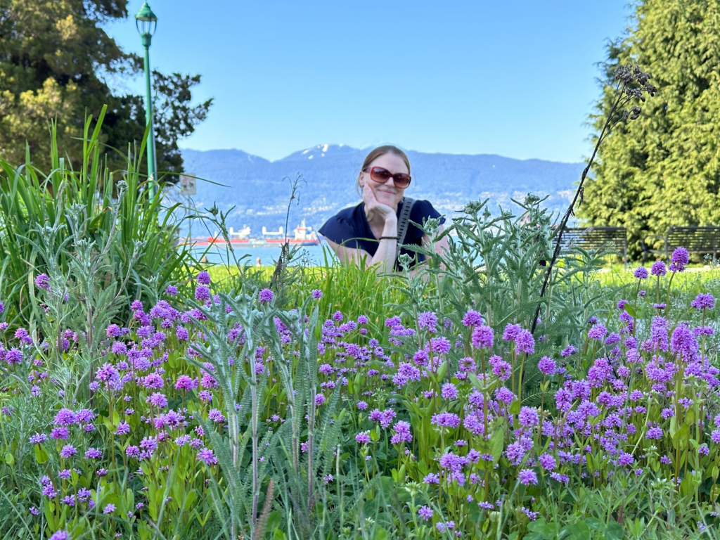 woman with sunglasses in front of a flowering meadow, with mountains and English Bay in background